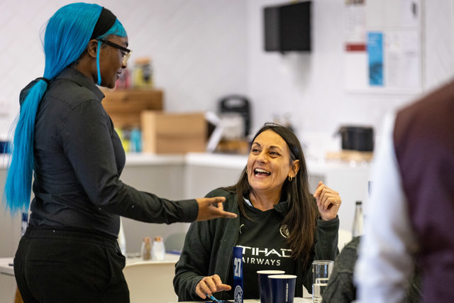 Two people engaging in a friendly conversation in an office setting. One person has bright blue hair and the other is smiling. Various cups are on the table.