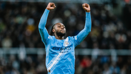 A Manchester City player wearing the team's blue jersey with arms raised in celebration. The jersey features the club crest and sponsorship logo.