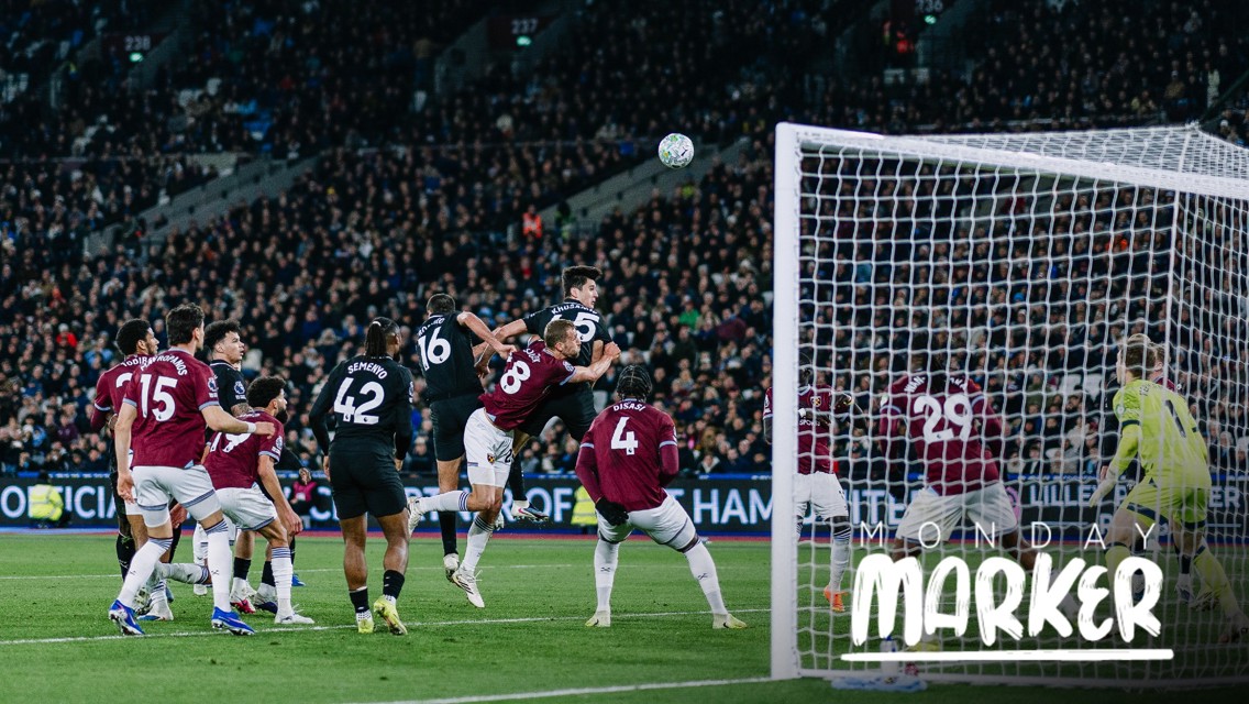 A group of soccer players from two opposing teams jump to head the ball during a match. The scene takes place near the goalpost with a crowded stadium in the background. Text 'Monday Marker' is visible.