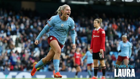 A football player in a light blue Manchester City jersey celebrates during a match at a stadium. Another player in red, possibly Manchester United, is visible in the background. Text says 'Thank You Laura'.