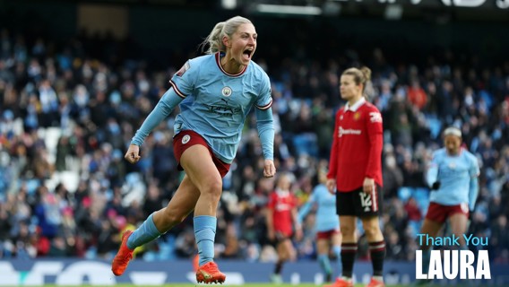 A football player in a light blue Manchester City jersey celebrates during a match at a stadium. Another player in red, possibly Manchester United, is visible in the background. Text says 'Thank You Laura'.