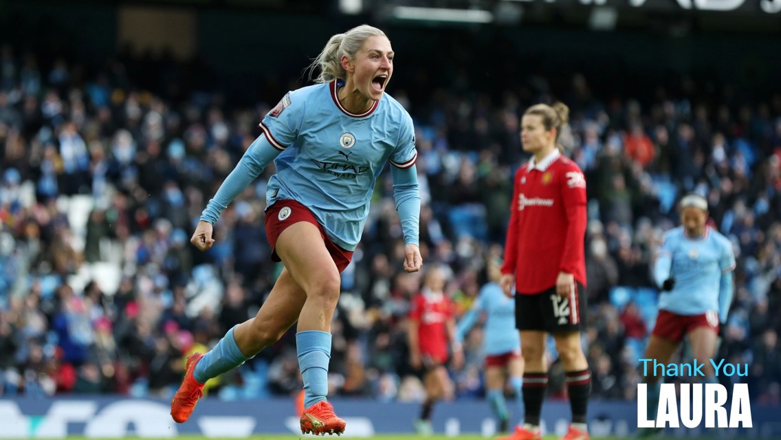 A football player in a light blue Manchester City jersey celebrates during a match at a stadium. Another player in red, possibly Manchester United, is visible in the background. Text says 'Thank You Laura'.