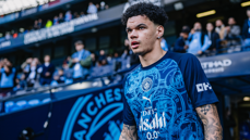 A person wearing a Manchester City training shirt in a stadium with a blurred face. The shirt features logos and sponsors, and the background includes a crowd and stadium signage.