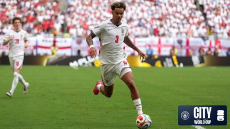 Soccer player wearing a white jersey with the number 3 on it running with the ball on the field during a World Cup match. The crowd is visible in the background with England flags and a logo for City at the World Cup in the corner.