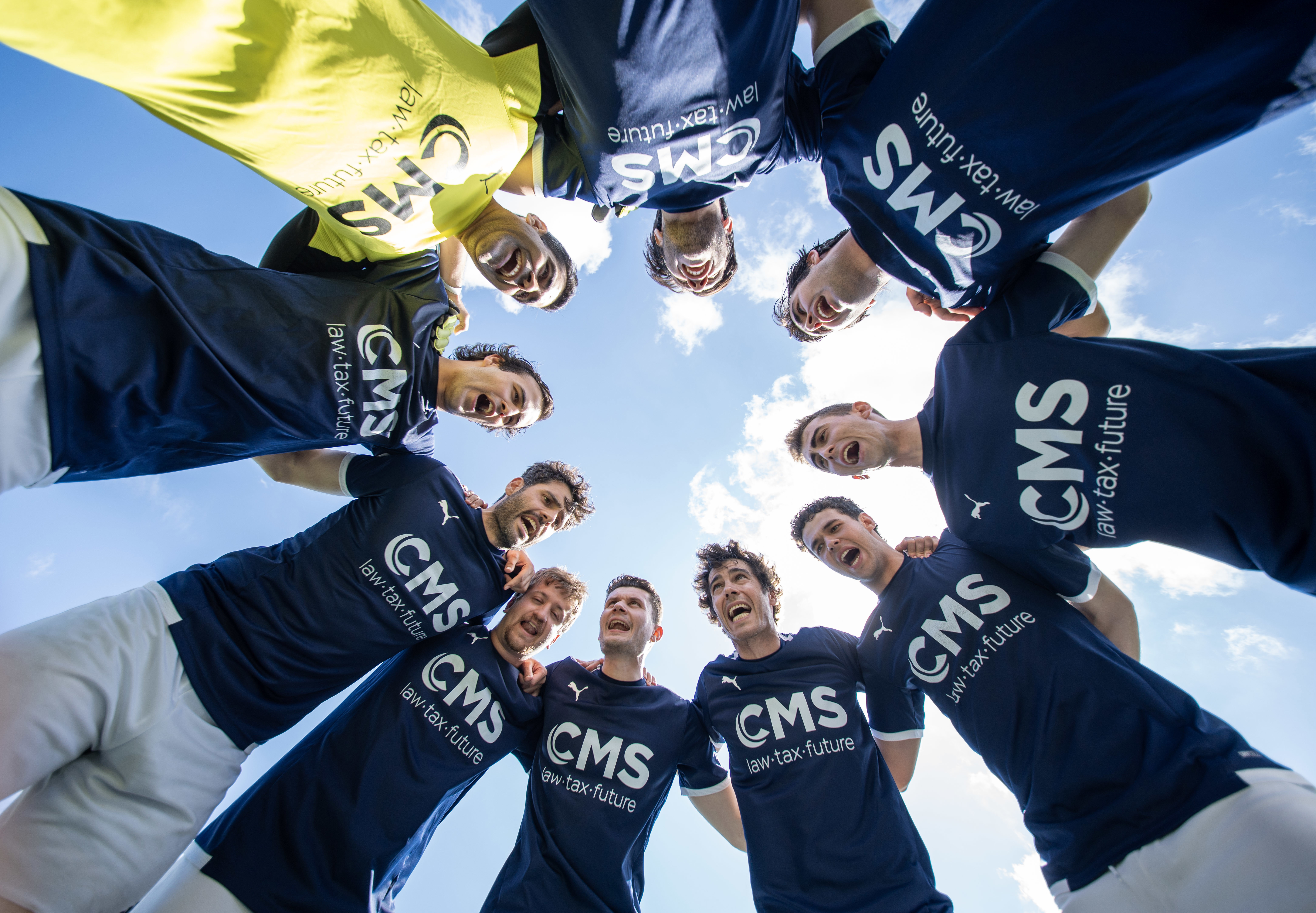 A group of soccer players in a circle with their arms around each other, looking down. The sky is visible in the background. They are wearing blue jerseys with 'CMS law.tax.future' written on them.