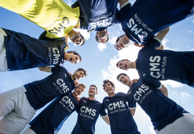 A group of soccer players in a circle with their arms around each other, looking down. The sky is visible in the background. They are wearing blue jerseys with 'CMS law.tax.future' written on them.