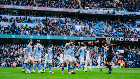 Manchester City players celebrating a goal at Etihad Stadium with a crowded audience in the background.