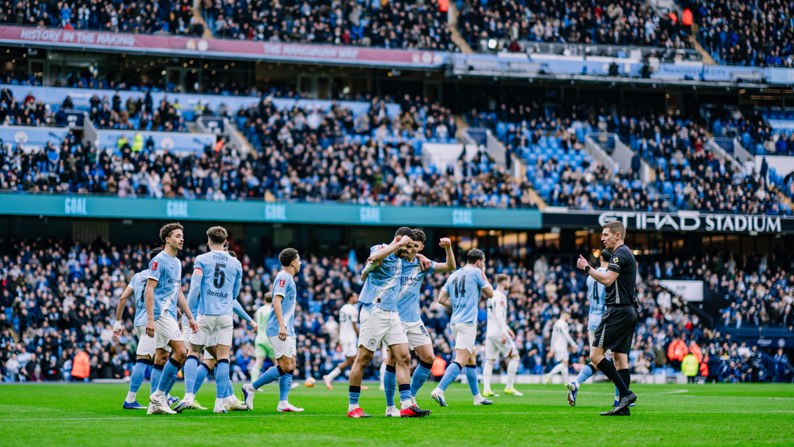 Manchester City players celebrating a goal at Etihad Stadium with a crowded audience in the background.