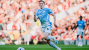 A football player in a light blue Manchester City kit is running on the field during a match, with a blurred crowd in the background.