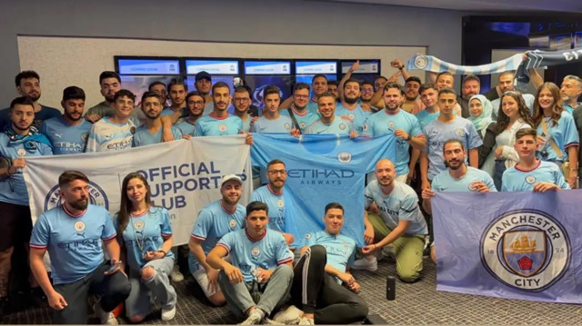 A group photo of Manchester City supporters wearing team jerseys, holding flags and banners with the Manchester City crest, in an indoor setting.