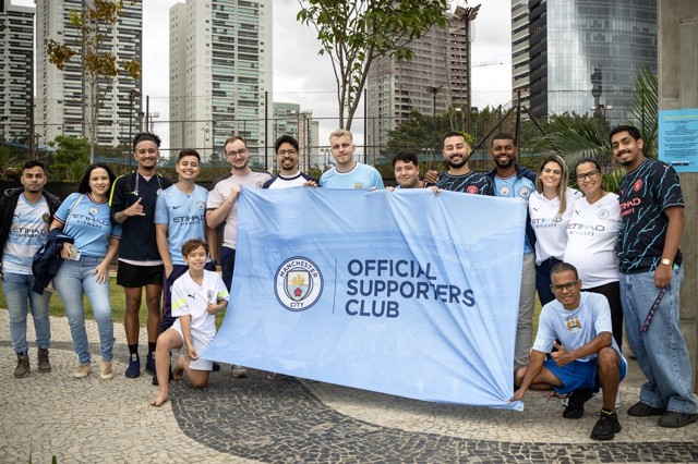 Group of Manchester City supporters holding an Official Supporters Club banner together with various Manchester City jerseys in front of a city background.