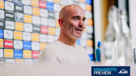 A football manager in a press conference setting, wearing a light sweater and holding a bottle of water. Background features sponsor logos.