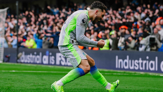 Soccer player in a gray and neon uniform celebrating a goal on the field with a crowd in the background.
