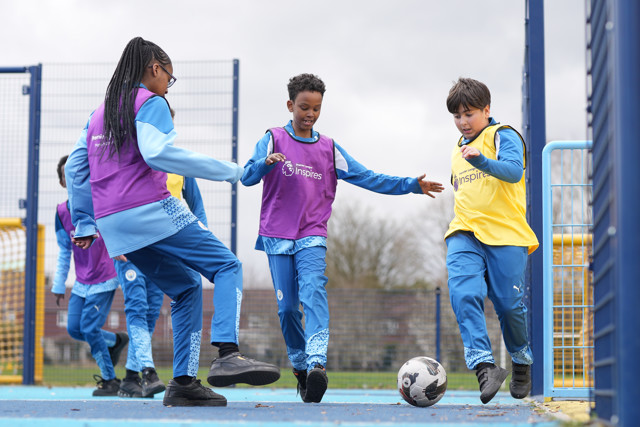 Children in blue sportswear engage in a football game on a playground, with two wearing yellow and purple bibs.