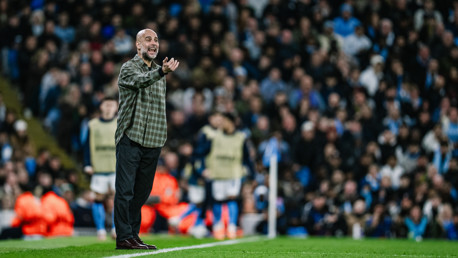 A manager is standing on the sideline of a football field, gesturing towards the pitch during a match. The background shows a crowded stadium with fans.