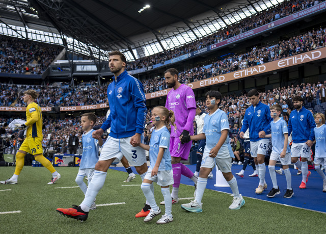 Football players and mascots walk onto the field at Etihad Stadium, wearing Manchester City colors with Etihad Airways branding.