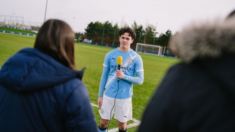 A Manchester City player in kit being interviewed on a football field, holding a microphone with two people in the foreground.