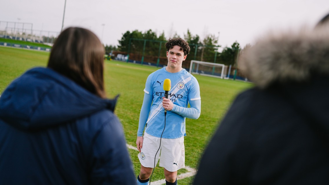 A Manchester City player in kit being interviewed on a football field, holding a microphone with two people in the foreground.
