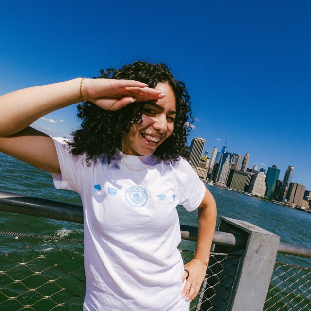 Person with curly hair wearing a white Manchester City themed shirt salutes at a waterfront with a city skyline in the background.
