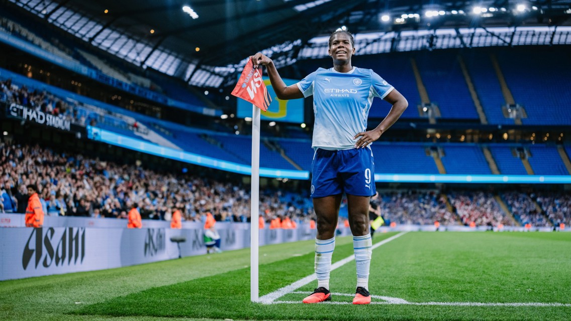 A Manchester City football player at the corner stand in Etihad Stadium, wearing the team's home kit, with a crowd in the background.