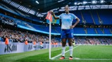 A Manchester City football player at the corner stand in Etihad Stadium, wearing the team's home kit, with a crowd in the background.