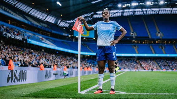 A Manchester City football player at the corner stand in Etihad Stadium, wearing the team's home kit, with a crowd in the background.