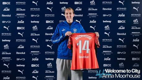Person holding a Manchester City jersey with 'Moorhouse' and number 14 on it. Manchester City branding in the background with various sponsor logos.