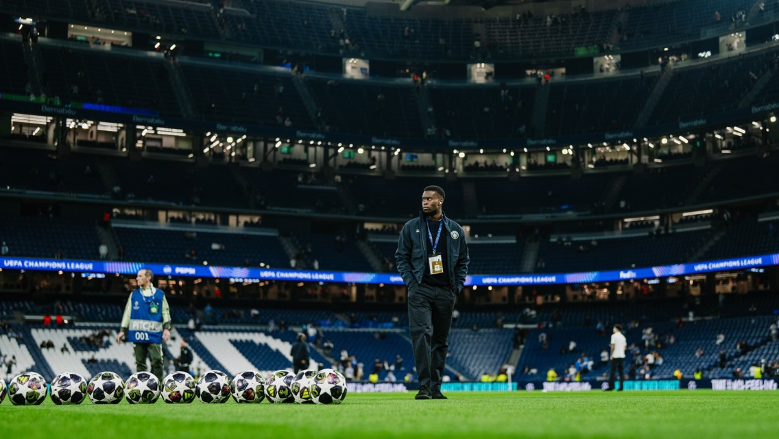 A person is standing on a football field with multiple soccer balls lined up in front of them, in an empty stadium with UEFA Champions League banners.