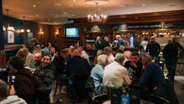 A pub setting with people seated at tables. The bar area is visible, displaying drink specials on a chalkboard. A TV shows a sports event.
