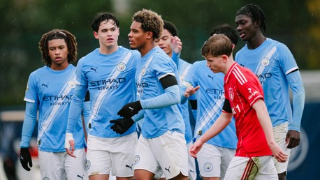 Manchester City youth players in blue kits celebrate on the pitch during a match against a team in red kits.