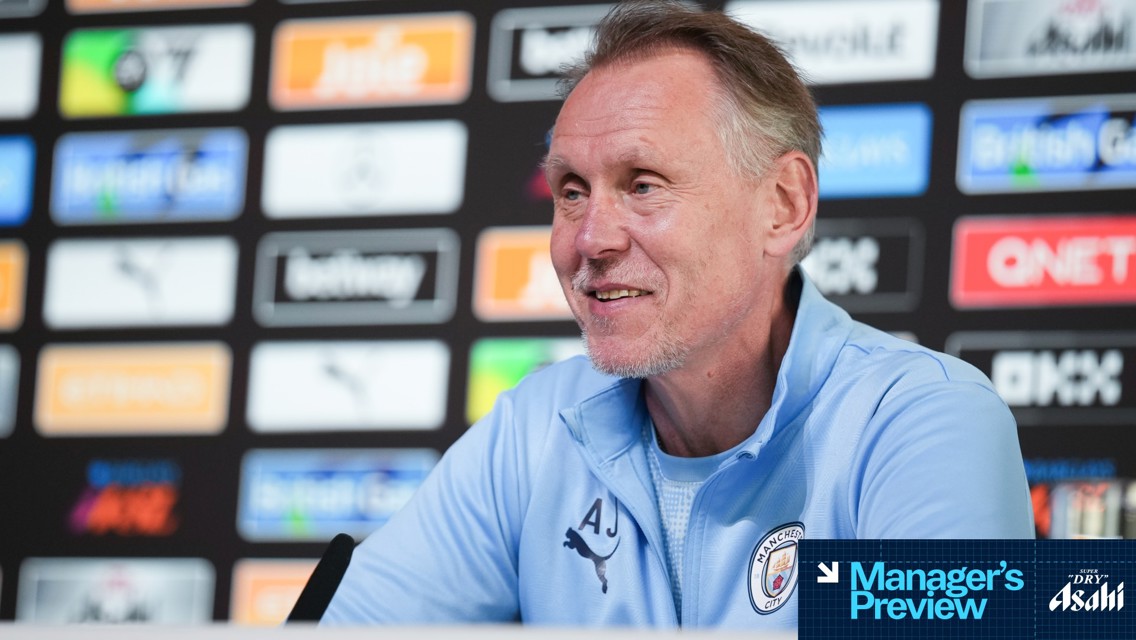 A Manchester City manager is speaking at a press conference, wearing a light blue Puma tracksuit with club logo. The background features various sponsor logos.