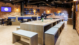 Modern lounge area with wooden tables and benches, blue cushions, exposed brick walls, and Manchester City decor including a crest on a TV screen.