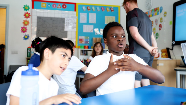 Students sitting at tables in a brightly decorated classroom with a teacher standing in the background.