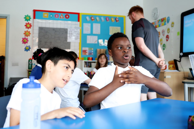 Students sitting at tables in a brightly decorated classroom with a teacher standing in the background.