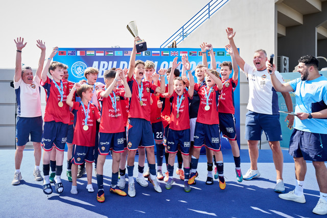 A youth soccer team celebrates a victory, holding a trophy. The team is wearing red jerseys and blue shorts, with medals around their necks. A banner with flags and a 'Manchester City' text is visible in the background.