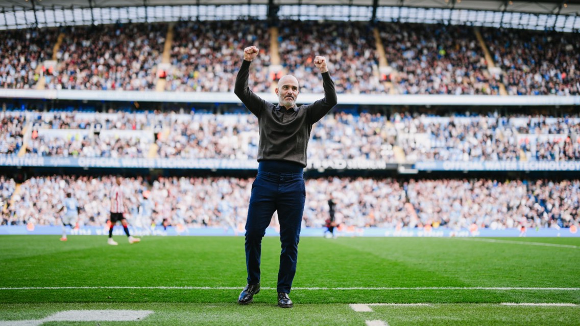 A person standing on a soccer field with their arms raised in celebration, surrounded by a full stadium crowd.