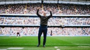 A person standing on a soccer field with their arms raised in celebration, surrounded by a full stadium crowd.