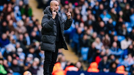 A football coach wearing a black coat gestures on the sideline in front of a blurred crowd during a match.