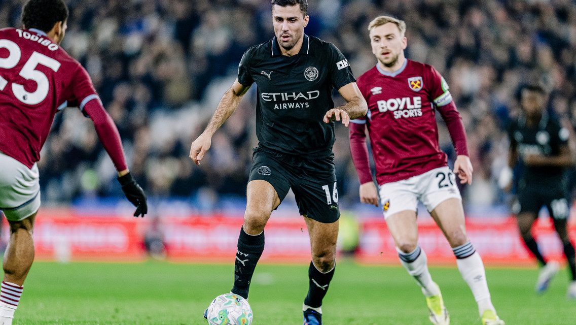 A football match with players from Manchester City and West Ham United. The Manchester City player controls the ball while surrounded by West Ham opponents.