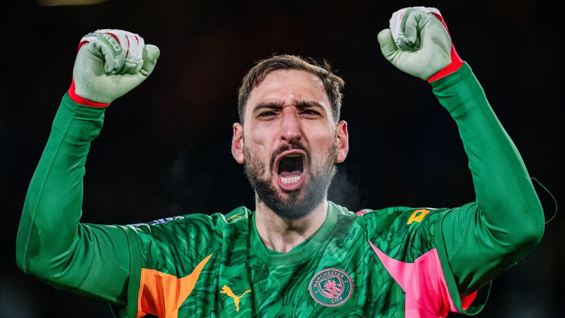 A football player in a Manchester City goalkeeper kit with arms raised in celebration. The kit features a green jersey with orange accents and club logo.