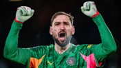 A football player in a Manchester City goalkeeper kit with arms raised in celebration. The kit features a green jersey with orange accents and club logo.