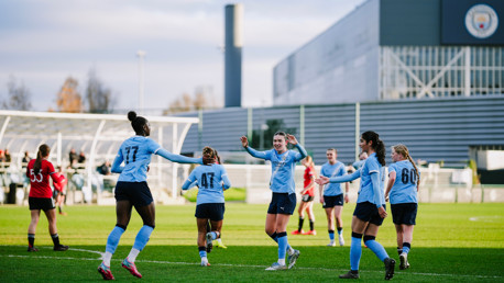 A women's soccer match featuring Manchester City players in light blue kits, including numbers 17, 41, 47, and 60. They appear to be celebrating a goal on a grass field with a Manchester City FC building in the background.