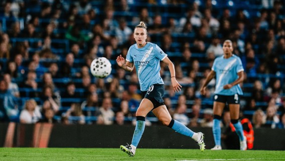 A female soccer player wearing a Manchester City kit is chasing a ball on the field during a match.