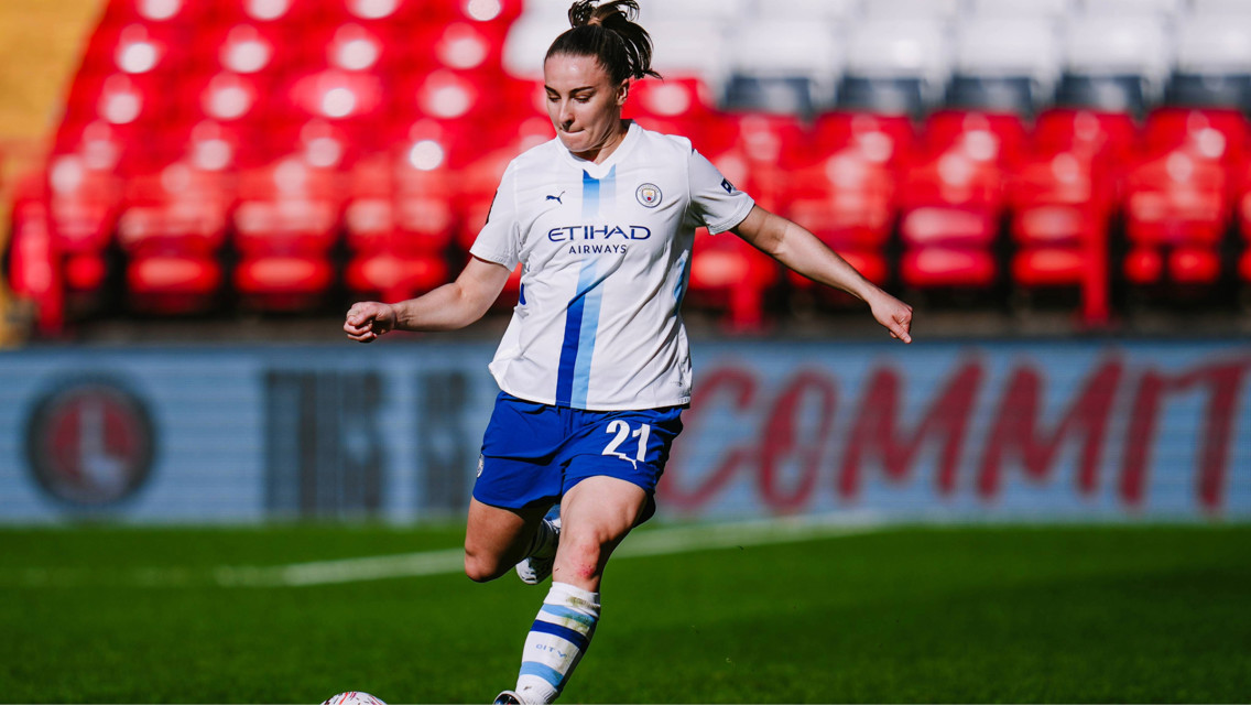 A female soccer player is kicking a ball on the field. She is wearing a Manchester City kit, with Etihad Airways as the sponsor.