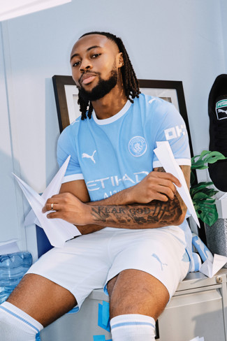A person in a Manchester City jersey holding paper planes, sitting on a table with plants and a Puma shoe in the background.