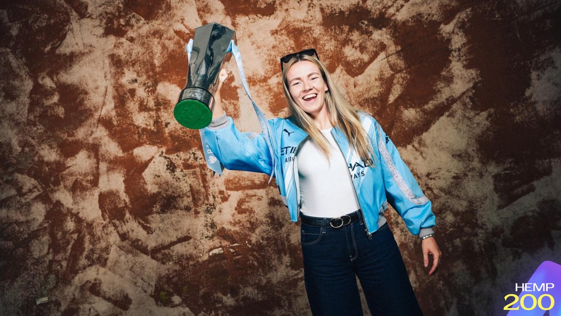 A woman in a light blue jacket holds a shiny trophy while smiling. The background is a textured brown surface. There is a digital graphic in the bottom right corner labeled 'HEMP 200'.