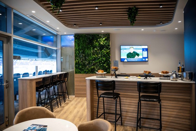 A modern lounge area with wooden decor, seating, a bar with pastries, a TV screen, and a window view of a stadium.