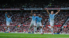 Four football players wearing light blue kits celebrate on a football field with a cheering crowd in the background. A banner above reads 'THE IMPOSSIBLE DREAM' and 'SIR ALEX 26 YEARS.'