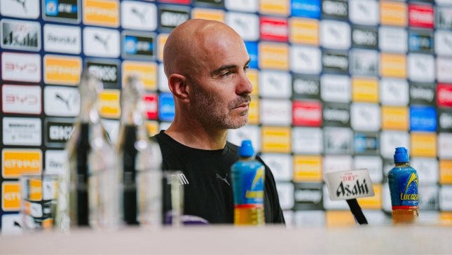 Person at a press conference stage with a backdrop of various logos, including Puma and Etisalat; Lucozade bottles and Asahi branding are visible on the table.