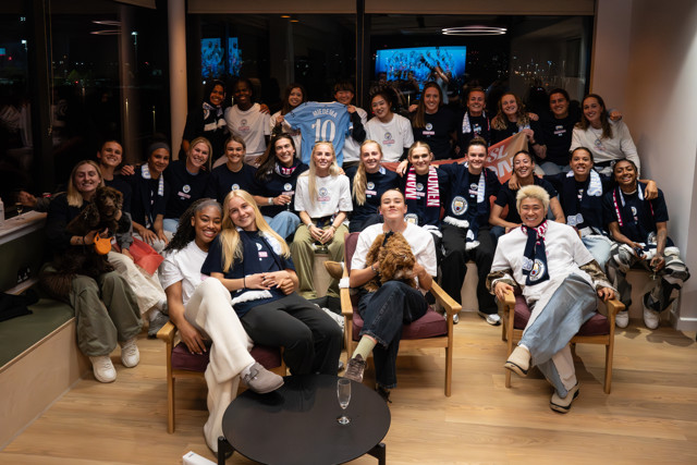 Large group wearing Manchester City gear, holding a shirt with 'Miedema 10'. There are scarves and a dog. The room has a cityscape view with lights outside.